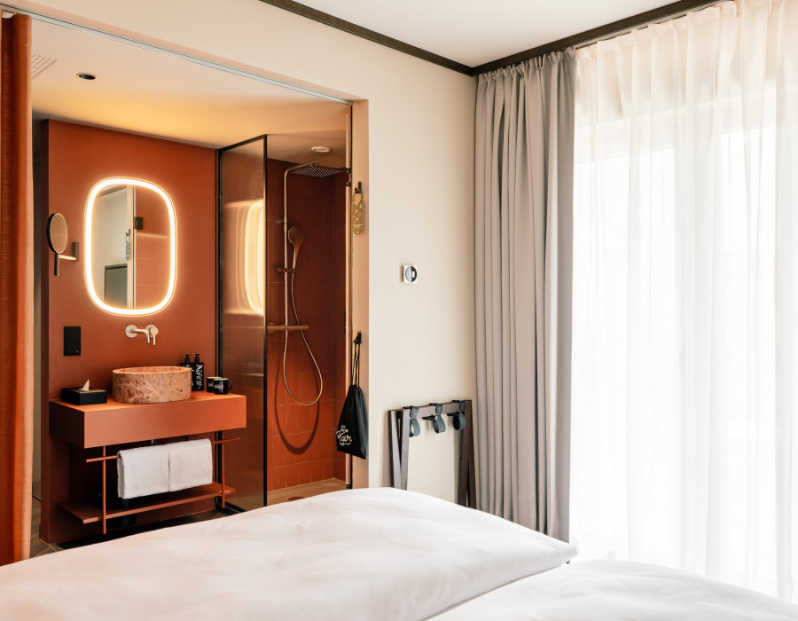Blick vom Bett in ein offenes Badezimmer mit Regendusche in einem Hotelzimmer mit Balkon