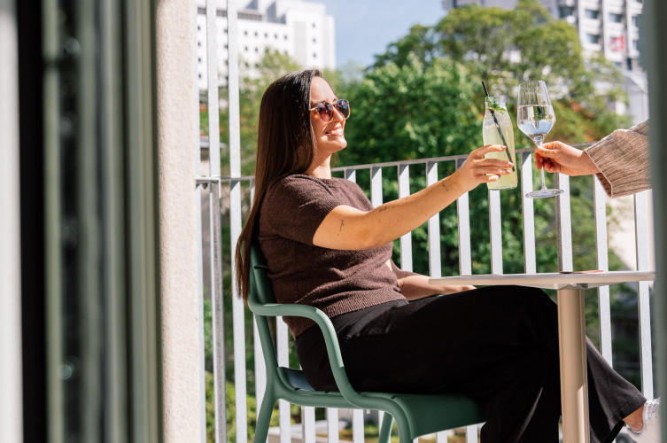 Eine Frau sitzt in der Sonne auf einem Balkon und stoßt mit einer anderen Person an 