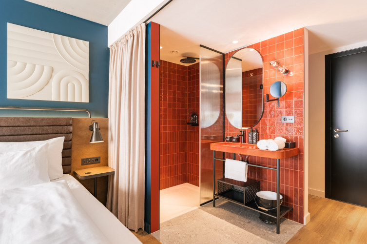 Offenes Badezimmer mit Regendusche, neben einem Doppelbett im Hotelzimmer beim Wiener Naschmarkt