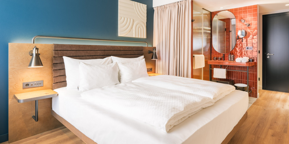 Doppelbett neben einem offenen Badezimmer mit Regendusche und Waschtisch im Hotel Schani Naschmarkt