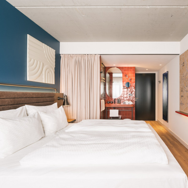 Doppelbett und offenes Badezimmer mit Waschtisch und Regendusche in einem Zimmer im Hotel Schani Naschmarkt