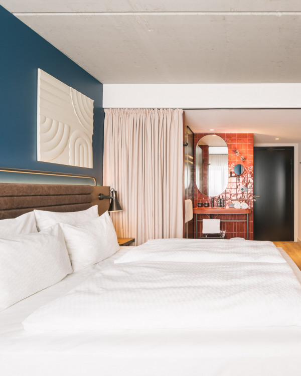 Doppelbett und offenes Badezimmer mit Waschtisch und Regendusche in einem Zimmer im Hotel Schani Naschmarkt