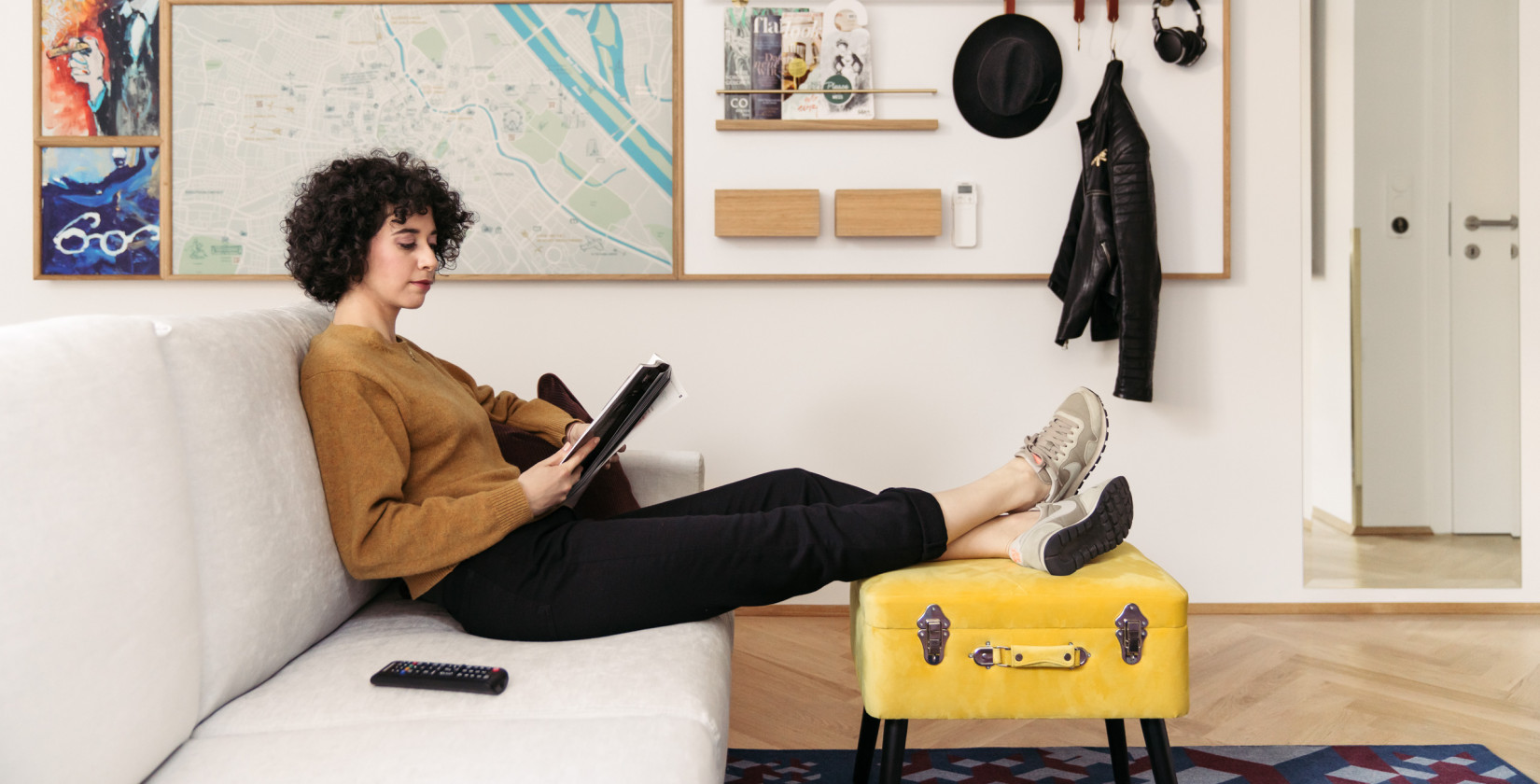 Frau sitzt auf einer Couch, liest und legt ihre Beine auf einen gelben Koffer