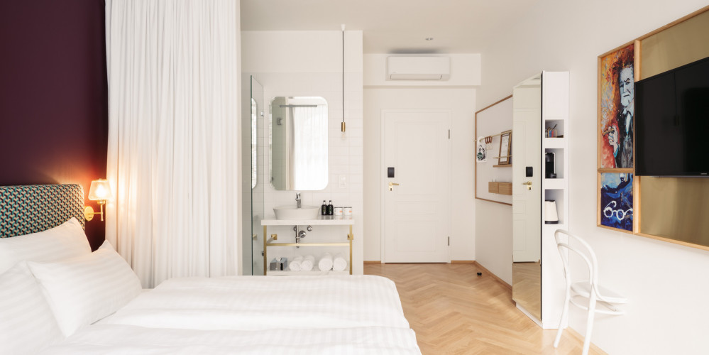 Light-filled room with a double bed against a violet wall opposite the TV set