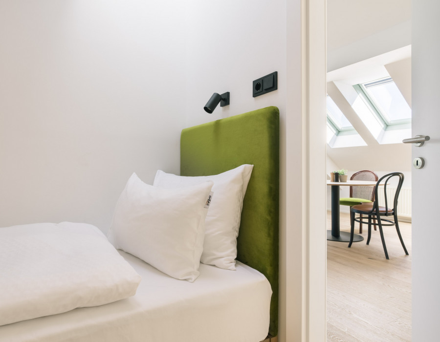 Einzelbett mit seitlich offener Tür und Blick auf den Essbereich mit Tisch & Stühlen im Hotelzimmer